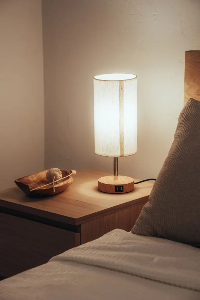 Table de chevet avec une lampe allumée à la lumière chaude, à côté d'un lit fait aux draps clairs.