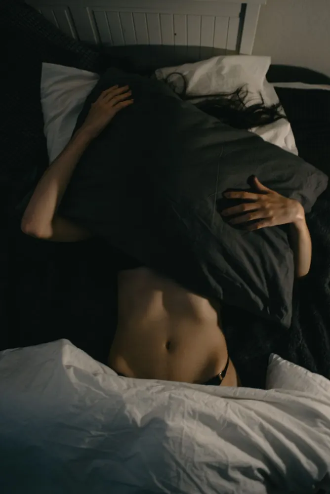 Femme allongée dans des draps sombres, le haut du corps et le visage cachés par un oreiller, seul le ventre est visible.