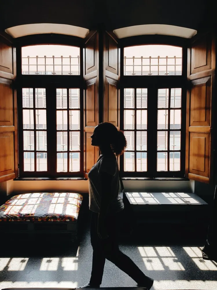 Silhouette sombre d'une femme qui marche devant deux grandes fenêtres lumineuses, à contre-jour.